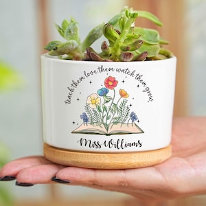 Könnte beinhalten: Weißer Keramik-Pflanztopf mit einer Sukkulentenpflanze. Der Pflanztopf hat einen Holzsockel und zeigt ein Blumendesign mit dem Text "teach them love them watch them grow" und "Miss Williams".