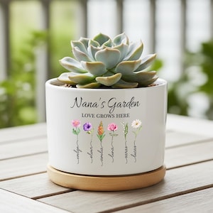 May include: White ceramic plant pot with a succulent. The pot has the text "Nana's Garden LOVE GROWS HERE" and floral illustrations with names. It sits on a wooden saucer.