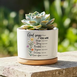 May include: A white ceramic plant pot with a succulent. The pot features the text "God says YOU ARE Unique, Strong, Beautiful, Forgiven, Precious, Never alone" and floral illustrations. It rests on a wooden base.