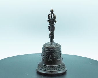 May include: A bronze-colored bell with intricate carvings and a decorative handle. The bell features detailed patterns and religious imagery, likely used for ceremonial purposes. The bell is displayed on a dark surface against a light background.