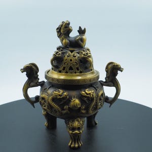 Könnte beinhalten: Bronze-Räuchergefäß mit aufwendigen Drachen- und Löwenmotiven. Der dreibeinige Räucherbehälter hat Griffe und einen dekorativen Deckel. Das Gesamtdesign ist verziert und detailliert, mit einer dunkelbronzenen Oberfläche.