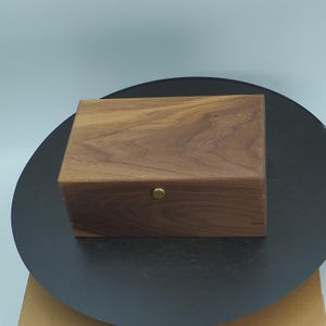 Könnte beinhalten: Eine rechteckige Holzkiste mit natürlicher Holzmaserung. Die Kiste hat einen messingfarbenen Knopf auf der Vorderseite und steht auf einer dunkelgrauen Oberfläche. Die Kiste scheint handgefertigt zu sein.