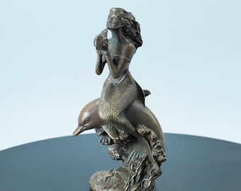 May include: Bronze sculpture of a mermaid riding a dolphin. The mermaid is holding a seashell and has flowing hair. The dolphin is leaping through stylized waves. The sculpture has a detailed, textured finish and is set against a light blue background.
