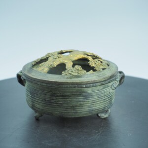 May include: A bronze incense burner with a tree design on the lid. The round container has a textured, ribbed exterior and small feet. The lid features a detailed, openwork tree design in a contrasting gold color.