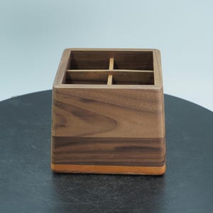 Puede incluir: Un organizador de escritorio de madera con cuatro compartimentos. El organizador es de madera marrón oscuro con una banda marrón claro y una base naranja. El diseño es simple y moderno, adecuado para el hogar o la oficina.