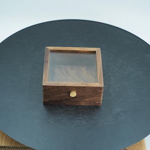 May include: A small, square wooden box with a clear glass top. The box has a drawer with a small, round, gold-colored knob. The wood grain is visible on the sides and front of the box.