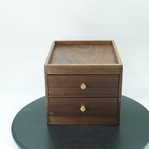 Può includere: Un piccolo cassettone in legno a due cassetti con finitura marrone scuro. I cassetti hanno pomelli rotondi color oro. La parte superiore del cassettone ha una superficie piana. Il cassettone è su una superficie nera.