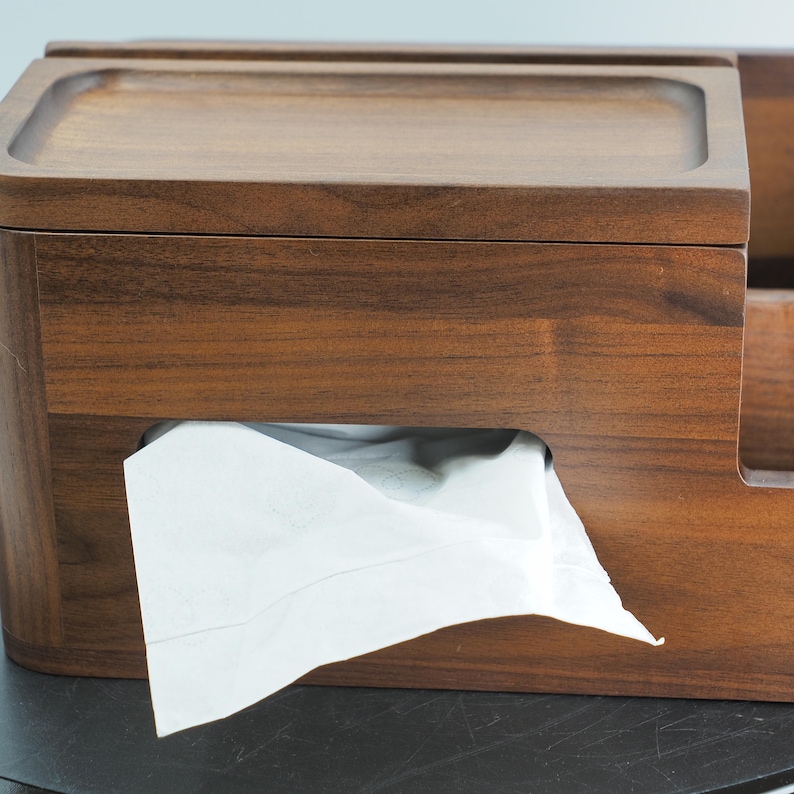 May include: A wooden tissue box with a dark brown finish. The box has a rectangular shape with a lid and a slot for dispensing tissues. White tissues are partially pulled out of the slot. The box is made of wood and has a smooth, polished surface.