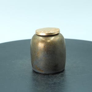 Puede incluir: Un pequeño tarro de cerámica hecho a mano con un esmalte metálico bronce y negro. El tarro tiene una forma redondeada y está rematado con una tapa plana de color dorado. El tarro se utiliza probablemente para guardar cosas o para decoración.