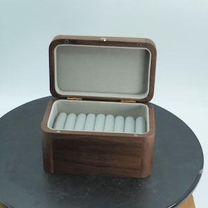 Könnte beinhalten: Eine rechteckige Schmuckschatulle aus Holz mit einem Klappdeckel, der sich öffnet und einen grauen Innenraum und Ringhalterungen freigibt. Die Schatulle ist aus dunkelbraunem Holz gefertigt und hat einen goldfarbenen Verschluss. Der Innenraum ist mit einem weichen, hellgrauen Stoff ausgekleidet.
