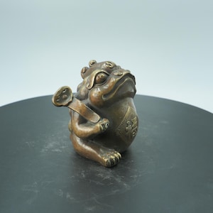 May include: A bronze frog figurine with a coin in its mouth and a pouch on its side. The frog is detailed with textured skin and sits upright. The pouch has an embossed design. The figurine is on a dark surface.