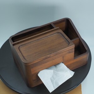 May include: A wooden tissue box with a built-in organizer. The box is made of dark brown wood and has a rectangular shape with rounded corners. A white tissue is pulled from the front. The organizer has multiple compartments.