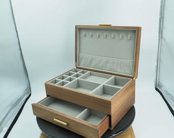 May include: A wooden jewelry box with a light brown finish, featuring multiple compartments and a pull-out drawer. The interior is lined with gray fabric. The box has a gold-colored latch and drawer pull.