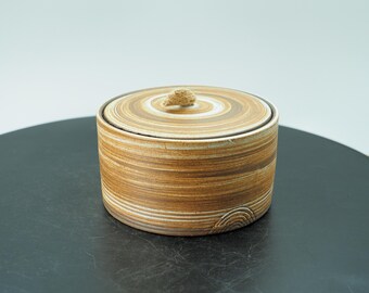 Puede incluir: Un recipiente redondo de cerámica con tapa, de estética natural y terrosa. El recipiente presenta bandas horizontales de tonos marrones y crema, con un diseño en espiral en la tapa. Un pequeño pomo texturizado sirve de asa.