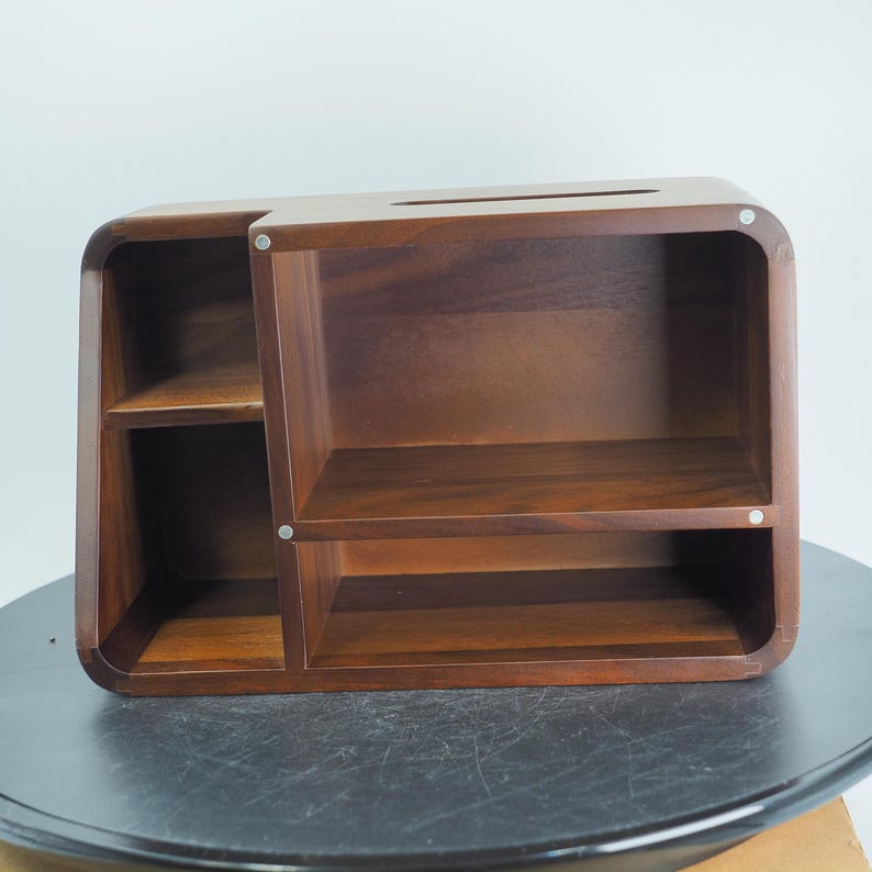 May include: A wooden desk organizer with a dark brown finish. The organizer has multiple compartments, including two shelves and a slot on top. The design features rounded corners and a modern aesthetic. The organizer is made of solid wood.