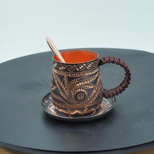 Puede incluir: Una taza de café decorativa con un platillo y una cuchara a juego. La taza es negra con intrincados diseños grabados en color cobre y un asa trenzada marrón. El interior de la taza es naranja. La cuchara descansa dentro de la taza.