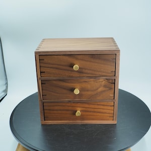 May include: A small, three-drawer wooden chest with a warm brown finish. The drawers have brass knobs and are set in a square frame. The chest is made of wood and has visible dovetail joints. The chest is sitting on a dark surface.