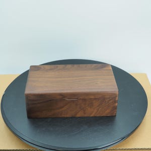 Puede incluir: Una caja rectangular de madera con tapa con bisagras. La caja está hecha de madera marrón oscuro con un patrón de vetas visible. La caja está sobre una superficie negra.