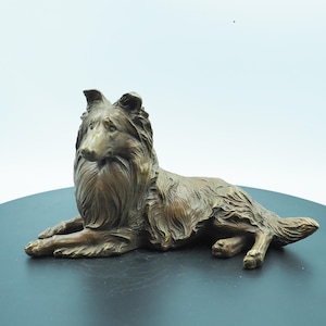 May include: A bronze sculpture of a Collie dog in a relaxed, lying-down pose. The dog has detailed fur texture and a long, flowing mane. The sculpture is set against a neutral background, highlighting the dog's form and features.