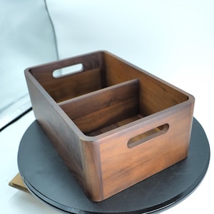 May include: A rectangular wooden storage box with a natural brown finish. The box is divided into two compartments and has handle cutouts. It is on a black surface.