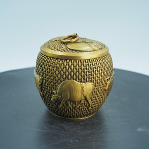 May include: A gold-coloured, round decorative box with a textured woven pattern. The box features embossed images of bison and a lid with a ring handle. The box is likely made of metal and has a vintage or antique aesthetic.