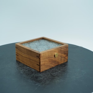 以下が含まれることがあります： 透明で質感のあるガラスの天板が付いた、小さな正方形の木製ボックス。ボックスは明るい茶色の木材で作られており、前面に真鍮製のノブが付いています。ボックスは暗い表面に置かれています。