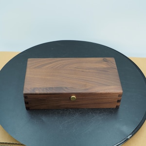 Może przedstawiać: Prostokątne drewniane pudełko z otwieraną pokrywą. Pudełko wykonane jest z ciemnobrązowego drewna z widocznym usłojeniem i mosiężnym pokrętłem z przodu. Pudełko jest zamknięte i stoi na ciemnej powierzchni.
