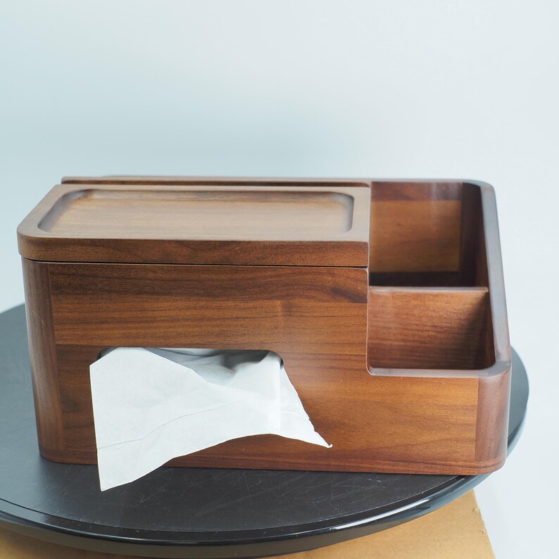 May include: A wooden tissue box with a built-in compartment. The box is made of dark brown wood and has a rectangular shape with rounded edges. White tissues are visible. The box sits on a black surface.