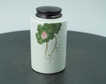 Puede incluir: Un tarro de cerámica blanco con tapa marrón oscuro. El tarro está decorado con una hoja de loto verde y una flor rosa. El tarro es cilíndrico y se apoya sobre una superficie oscura.
