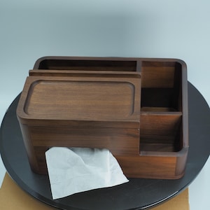 May include: A dark brown wooden tissue box with a rectangular shape. The box has a sliding top and multiple compartments for storage. White tissue paper is visible, pulled from the dispenser. The box is on a dark surface.