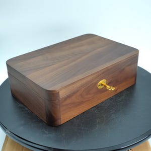 May include: A rectangular wooden box in a dark brown finish. The box features rounded corners and a brass keyhole with a small key inserted. The wood grain is visible on the lid and sides, suggesting a handcrafted item.