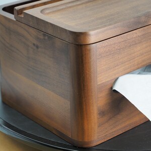 May include: A wooden tissue box with a dark brown finish. The box has a rectangular shape with rounded corners and a lid. White tissue paper is visible from the dispensing slot. The box is sitting on a black surface.