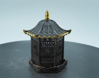 Puede incluir: Un pequeño objeto decorativo en forma de pagoda, de color marrón oscuro, con adornos dorados en el techo y un pináculo. La pagoda tiene intrincados patrones calados en sus lados y se asienta sobre una pequeña base cuadrada. El diseño recuerda a la arquitectura asiática.