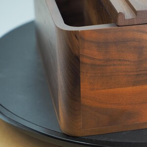 May include: A close-up of a wooden box with a rich, dark brown finish. The box has a rectangular shape with rounded corners and a smooth, polished surface. The wood grain is visible, adding to its natural aesthetic.