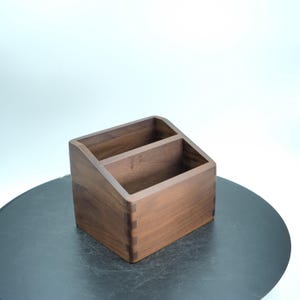 Pode incluir: Um organizador de mesa de madeira com dois compartimentos. O organizador é feito de madeira marrom escura e tem um topo inclinado. As juntas são visíveis, mostrando o artesanato. O organizador está sobre uma superfície escura.