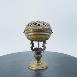May include: An antique brass incense burner with a spherical body and ornate lid. The lid has intricate cut-out designs, and the burner sits on a decorative stand. The design is symmetrical and detailed, with a warm, aged patina.