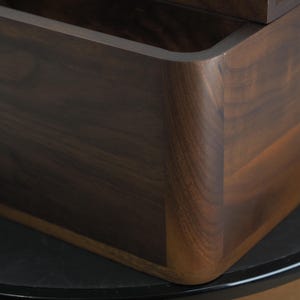 May include: Close-up of a dark brown wooden box with rounded corners. The wood grain is visible, and the box appears to be well-crafted. The box is sitting on a black surface.