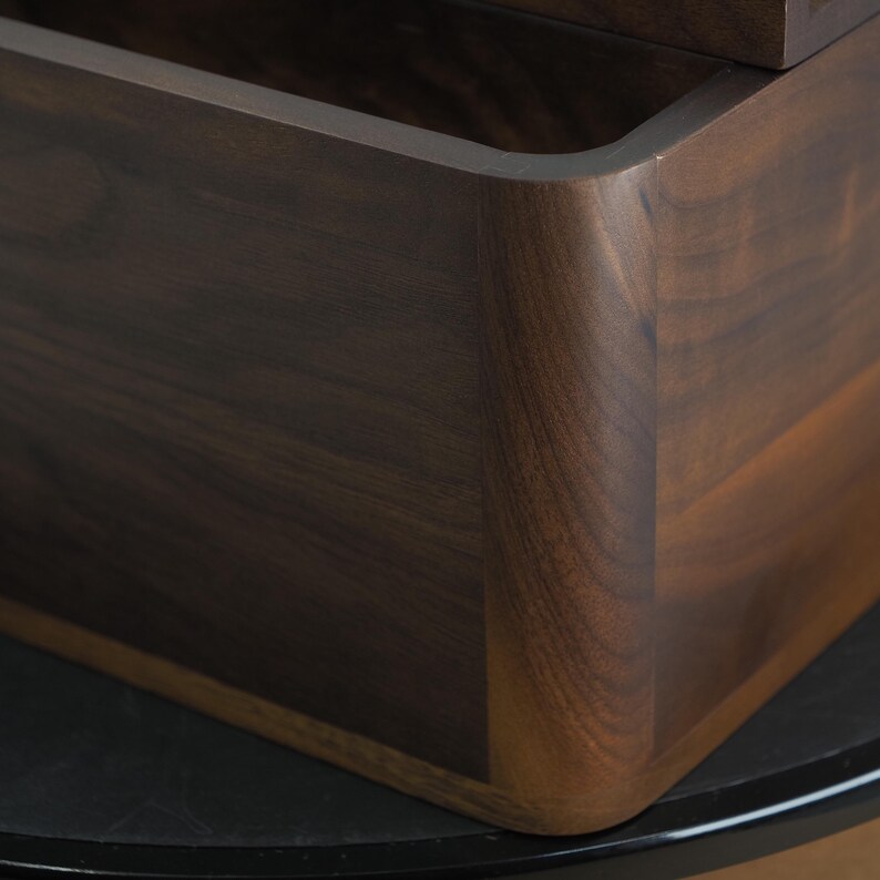 May include: Close-up of a dark brown wooden box with rounded corners. The wood grain is visible, and the box appears to be well-crafted. The box is sitting on a black surface.