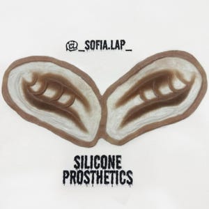 Puede incluir: Dos prótesis de oreja de silicona marrón con un diseño detallado, sobre un fondo blanco. El texto "SILICONE PROSTHETICS" está impreso en negro debajo. El nombre del artista, "_SOFIA.LAP_," está impreso en negro arriba.