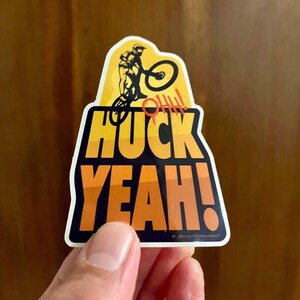 May include: A sticker featuring a mountain biker in yellow and black. The sticker reads "HUCK YEAH!" in orange and black, with "HUCK" above "YEAH!". The word "OHH!!!" is in red. The sticker is held by a hand.
