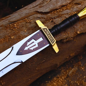 May include: A detailed image of a sword with a silver blade and a brown and gold hilt. The hilt features a black leather grip and gold accents. The blade has a brown emblem with a trident symbol. The sword rests on a wooden surface.