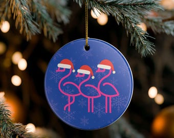 Może przedstawiać: Okrągła, niebieska ozdoba choinkowa z trzema różowymi ilustracjami flamingów w czapkach Świętego Mikołaja. Ozdoba ma złotą nić do zawieszenia i jest ozdobiona białymi płatkami śniegu.