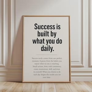 Könnte beinhalten: Ein rechteckiges Wandbild in einem hellen Holzrahmen mit schwarzem Text auf cremefarbenem Hintergrund. Die große Überschrift lautet Success is built by what you do daily, gefolgt von einem Absatz mit kleinerem Text über Gewohnheiten.