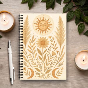 Puede incluir: Cuaderno de espiral beige con un diseño de sol, luna y flores en tonos marrón y dorado. Un bolígrafo blanco y dos velas encendidas sobre una superficie de madera, con hojas verdes alrededor.
