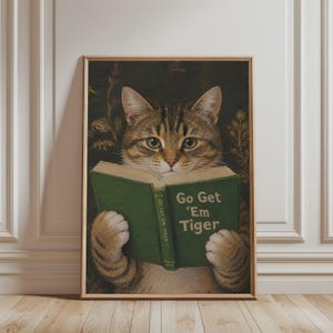 Op de afbeelding: Een ingelijste wandkunstprint met een bruine cyperse kat die een open groen boek vasthoudt. De boekomslag toont de tekst Go Get 'Em Tiger in witte letters. De kat heeft groene ogen en staat tegen een donkere, bloemrijke achtergrond.