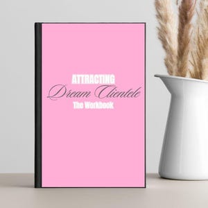 Puede incluir: Un cuaderno rosa con el título "ATTRACTING Dream Clientele The Workbook" en texto blanco. El cuaderno tiene un lomo negro y está junto a una jarra blanca con hierba decorativa seca.
