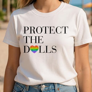 Può includere: T-shirt bianca con la scritta "PROTECT THE DOLLS" in lettere maiuscole nere. La parola "DOLLS" presenta un design a cuore arcobaleno. La maglietta è indossata all'aperto con palme e cielo blu sullo sfondo.