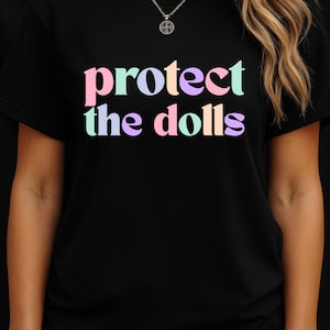 Può includere: T-shirt nero con la scritta "protect the dolls" in colori pastello. Il testo è in un carattere giocoso e arrotondato. La maglietta è un classico girocollo.