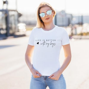Può includere: T-shirt bianco girocollo con la scritta "LIFE IS BETTER with my boys" in corsivo nero. La maglietta presenta un piccolo cuore nero. La persona nell'immagine indossa jeans blu e occhiali da sole.