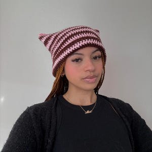 Könnte beinhalten: Handgefertigte Häkelmütze mit Katzenohren in braunen und rosa Streifen. Der Hut ist ein einzigartiges Accessoire, perfekt, um jedem Outfit einen Hauch von Laune zu verleihen. Eine stilvolle und gemütliche Wahl für kühleres Wetter.
